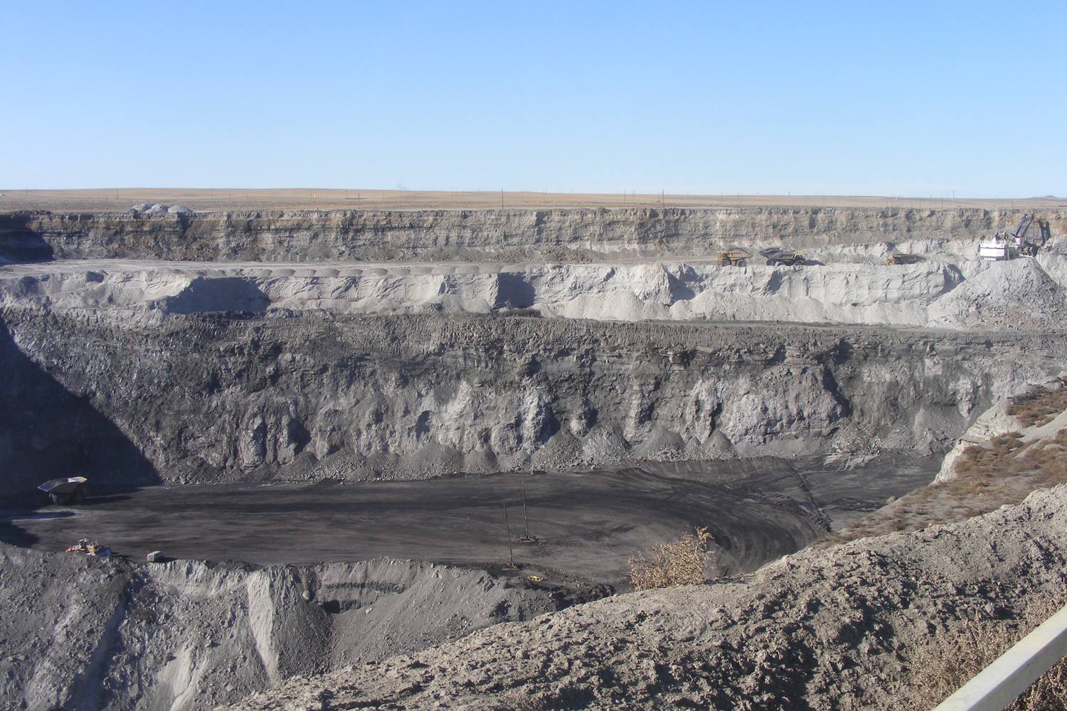 A black coal seam exposed at the bottom of a wide open pit mine below pale benches of overburden under a clear blue sky.