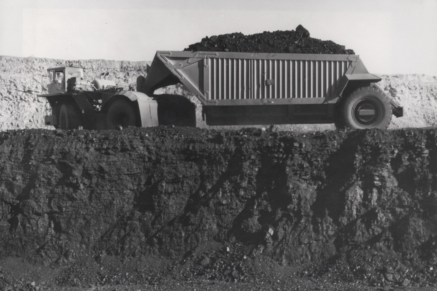 A loaded coal hauler parked on a bench of pale rock above a dark seam in a black and white view of an open pit mine.