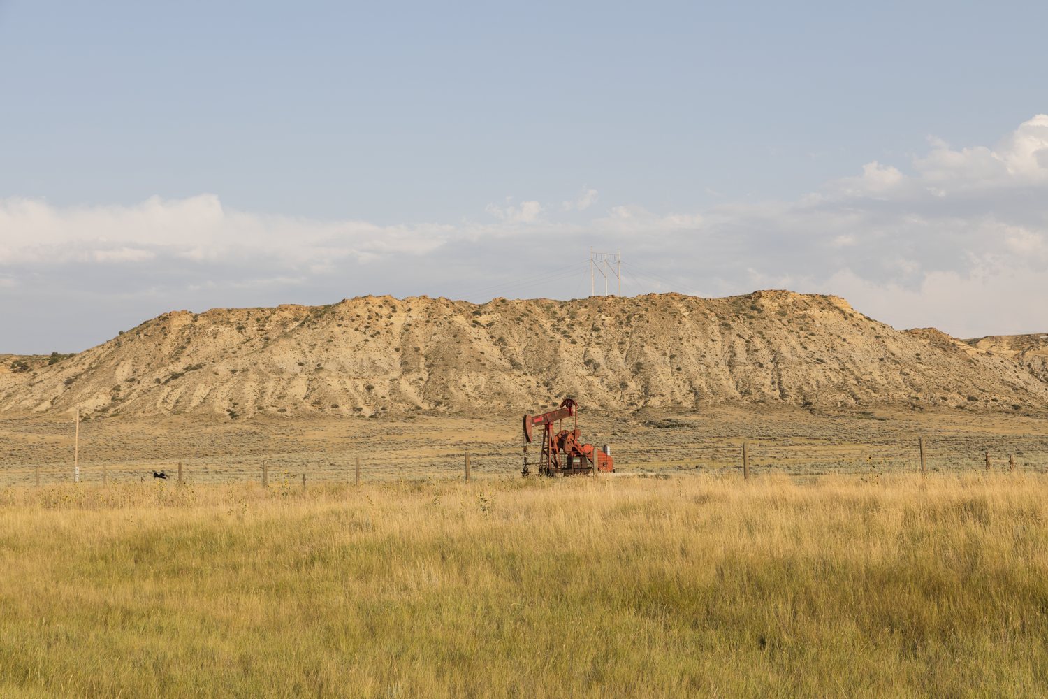 A single red pumpjack standing in dry grass and sagebrush below a low eroded bluff under a hazy summer sky.