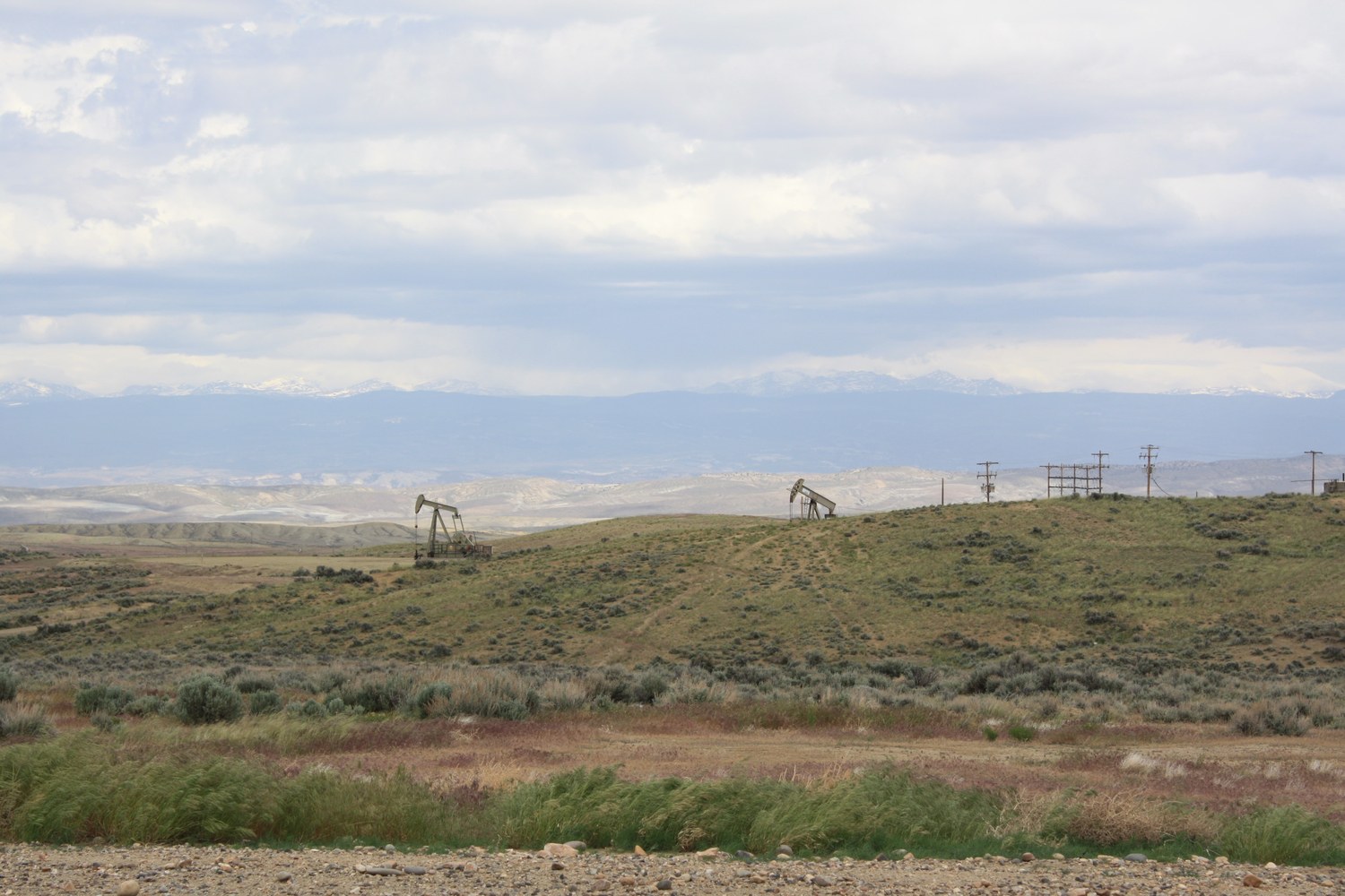 Two pumpjacks working a sagebrush oil field on rolling hills with a row of power poles and a line of snowy mountains under grey cloud.