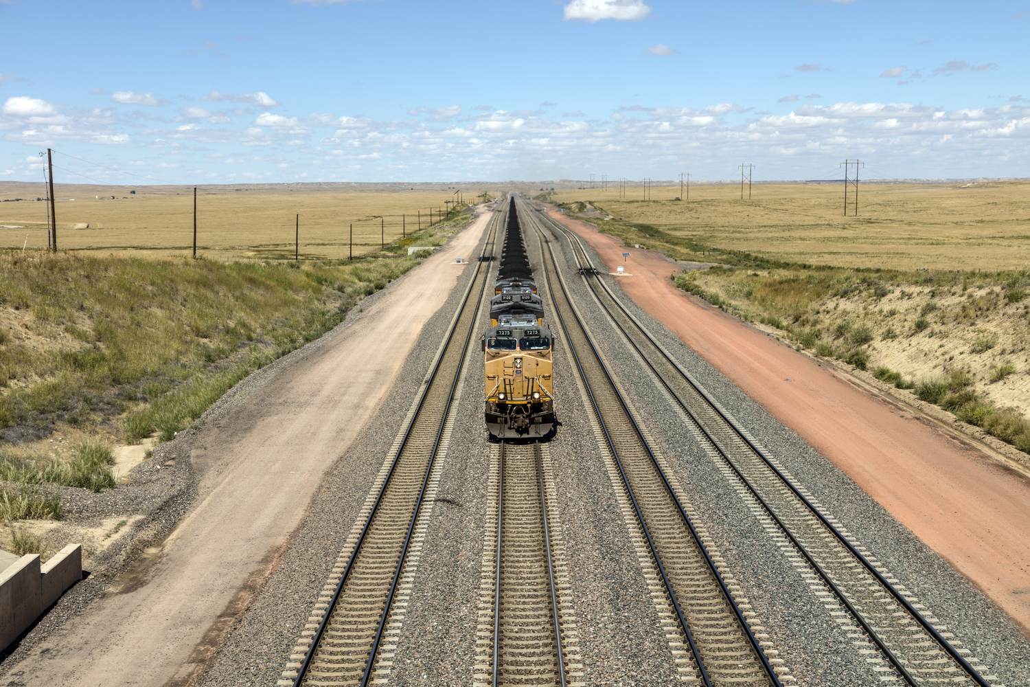 A yellow locomotive leading a long loaded coal train down straight triple track through flat dry grassland under a wide sky.